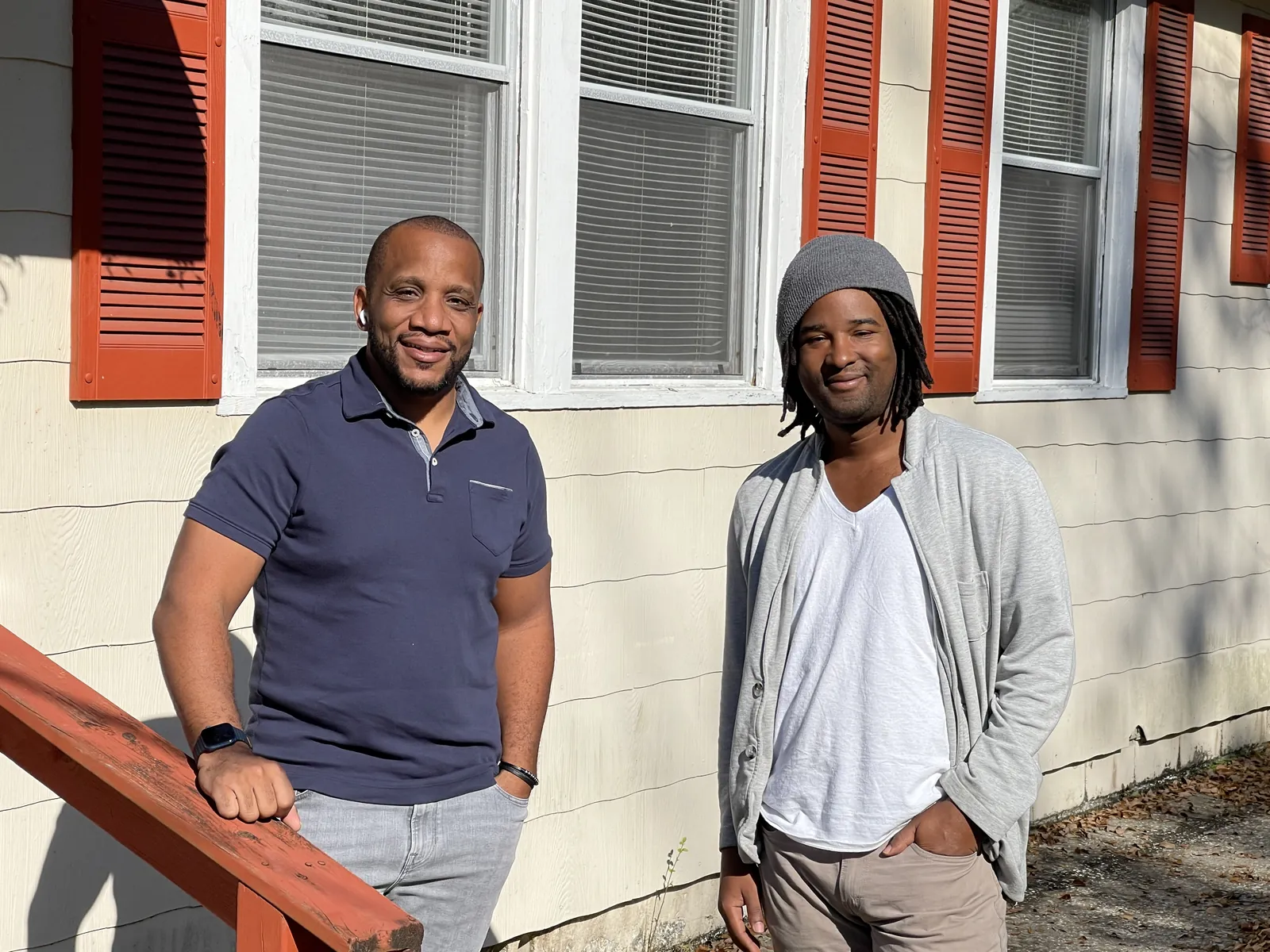 Two men standing outside an older single story Florida home with red shutters