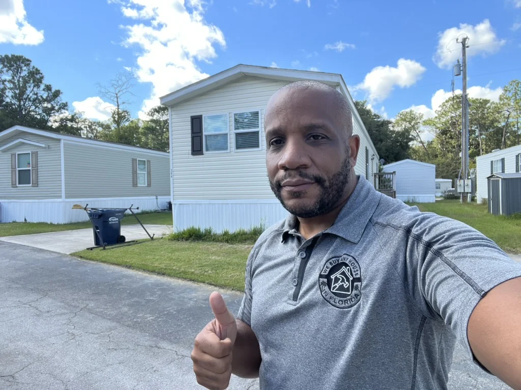 Chris Moore giving a thumbs up in a We Buy Any House In Florida polo shirt in front of a single story home