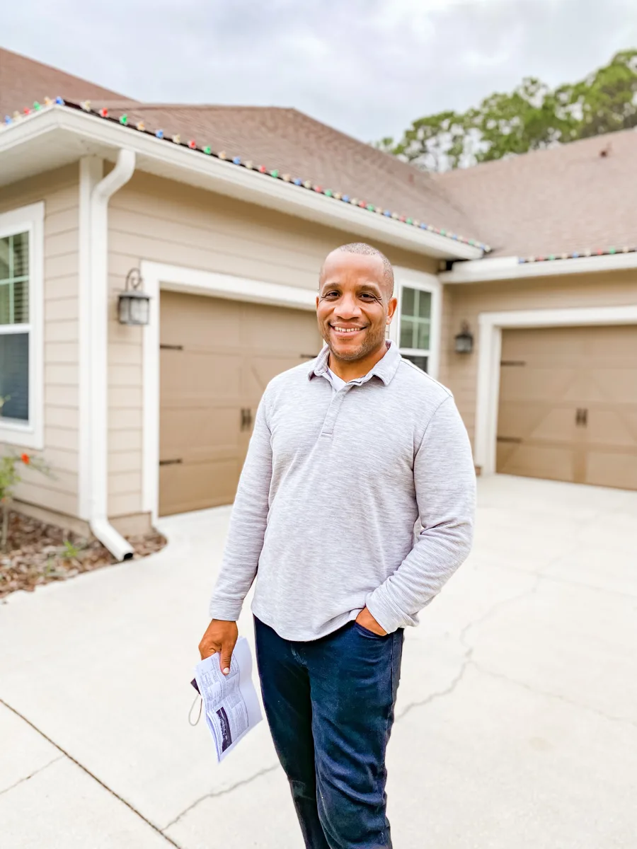 Chris Moore smiling in front of a two car garage on a single story home, holding a few papers