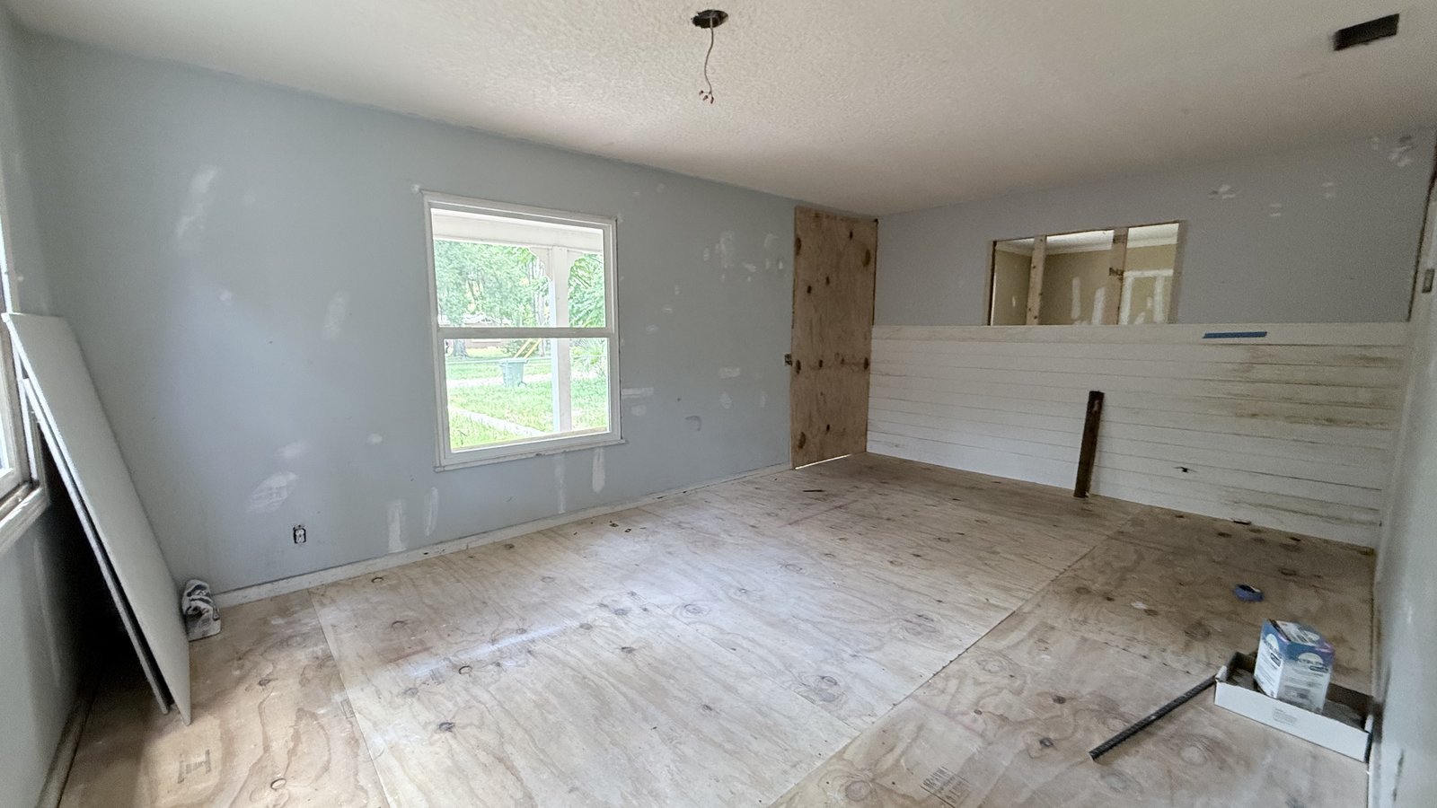 An empty room under renovation with a bare plywood floor, patched blue-gray walls, a window, and white wall paneling along one side.